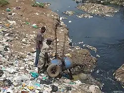 Two men on a trash strewn river bank emptying a barrel of human waste into the river