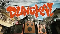 A photo of numerous loudspeakers stacked on top of one another; the song's title, "Dungka!", is superimposed on the photo in bold orange font