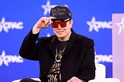 Elon Musk wearing an unbuttoned blazer with a gold chain and graphic t-shirt underneath, a pair of mirrored sunglasses, and a "Dark MAGA" hat – i.e. a black hat with "Make America Great Again" written in a gothic font.