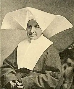 An older white woman in the habit of a religious sister, with a wide white wimple. She is holding rosary beads.
