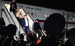Vadim Krasiko in a tracksuit and cap and three other men descend from an airplane labeled ТҮ-204-300. The airplane and the stairs are labeled "Russia" in Russian and with the three colors of the Russian flag. The heads of two soldiers with guns mounted with bayonets partially block the view.