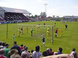 A football game in progress between F.C. United in red and Leigh Railway Mechanics Institute in yellow. The F.C. United crowd watch on.