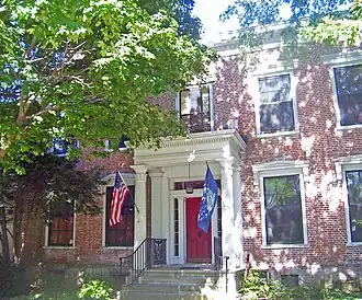 A brick building with a red door behind a small columned porch with the U.S. and New York flags behind a large tree on the left