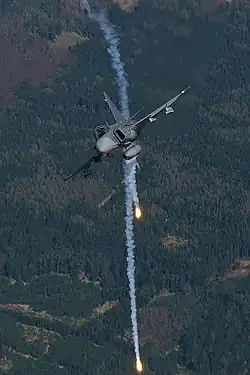 Head on view of fighter jet banking right while releasing flares against a background of green woodland