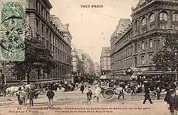 A sepia photograph looking down a built-up street, with the tramway to the right. A small crowd waits to board the tram.