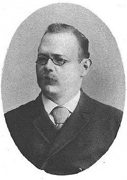 A black and white portrait of an older man with glasses, a mustache, and a receding hairline, wearing a dark suit.
