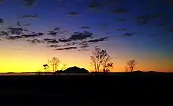 Small, pointed mountain on the horizon, foregrounded by scattered bare trees, silhouetted against the sky at sunset