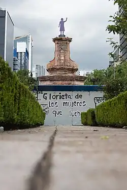 The sculpture depicts a woman with her left hand raised. It stands on a pedestal surrounded by police protective fencing, which bears the phrase "Glorieta de las Mujeres que Luchan".