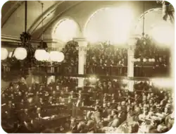 A photograph showing a large number of men seated on semi-circular tiers in a vaulted chamber as a large crowd looks on from an arcaded balcony
