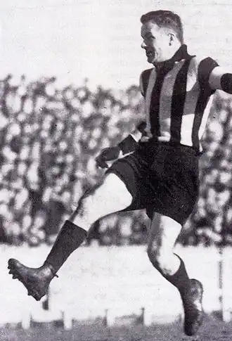 A dark-haired footballer in a kicking motion wearing a long-sleeve black-and-white vertically striped guernsey, dark shorts and football boots