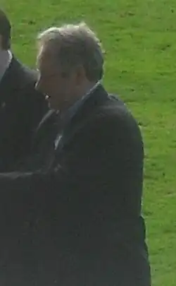 A man standing on a grass football pitch, wearing a black suit.