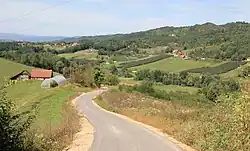 Picture of a countryside taken from the middle of a small road. There are hills covered in trees on the right and some farm structures in the distance and to the left.
