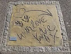 Handprints in a sand, with a heart-shaped and arrow drawings and written words "With love Kylie x".