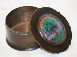 Colour photograph of a circular copper box with lid, depicting a tree in enamel, propped against the body