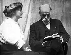 A white woman and a white man, seated. The woman, in profile, has her hair arranged in a bouffant updo, and is wearing a white blouse with puffy sleeves. The man, older, is bald, wears a mustache and a dark suit, and has his head bent to read a book.