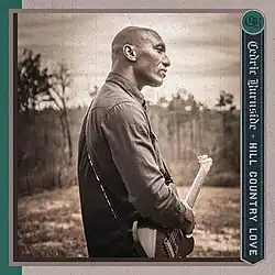 Cedric Burnside, standing outside in a rural area with an electric guitar
