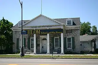 A light brown stone building with a pointed roof, green shutters and white columns on the front. In front of it are a sidewalk, a blue marker with gold writing, and a portion of street. Black letters above the front columns read "Holland Land Office Museum", which is also on a banner across the front.