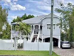 A long shot of a high-set Queenslander residential house made of timber with a corrugated iron roof. An external staircase leads to a veranda on the upper level of the house, and a white picket fence sits at the front of the property.