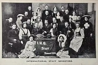 Postcard showing the International Staff Songsters wearing early 20th century Salvation Army uniforms, around the time of the 1904 International Congress in London. A banner in front of the group reads 'I.H.Q. Staff Songsters' and a caption underneath says 'International Staff Songsters'.