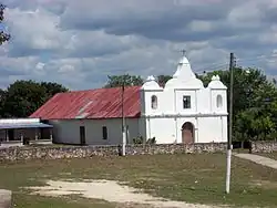 An open field with patchy, worn grass extending towards a low church with a simple white facade. The facade has three short spires, with the middle spire taller than the flanking spires. The central spire supports a cross. The shorter spires sit above arched openings, a bell is visible in the left-hand arch. An arched wooden door is set into the facade; a square window is above it. The main body of the church only stands about half the height of the facade and extends back to the left. The wall is bare except for a smaller doorway and a small window. The roof is rusty corrugated metal. A lower building with a shaded balcony extends to the left of the church. Two poles support a power line running across the field to the church. The base of each pole is painted white. A line of trees runs across behind the church.