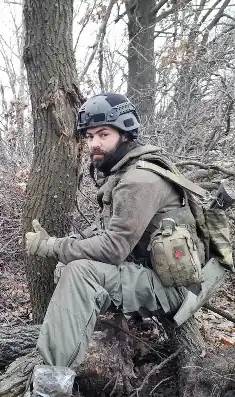 Mangushev in Olive green military clothing and a ballistic helmet, with a rifle over his shoulder and a first aid kit on his hip giving a thumbs-up signal towards the photographer