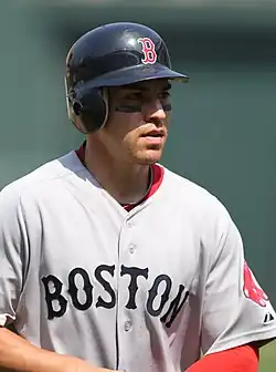 A young man in a grey baseball jersey wearing a batting helmet with eyeblack under his eyes.