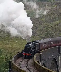 A steam train approaching the camera and emitting smoke while crossing a raised, arched bridge.