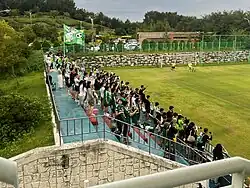 Football fans wearing green and white Jeonbuk Hyundai jerseys, and holding various flags and banners, cheer from the stands of a small football pitch.