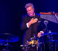 John Robinson standing behind the drums, wearing a black suit, hand on his heart to acknowledge applause