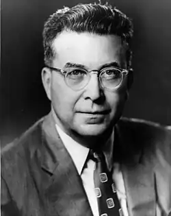 Middle aged, clean-shaven man with tousled dark hair, round spectacles and a suit