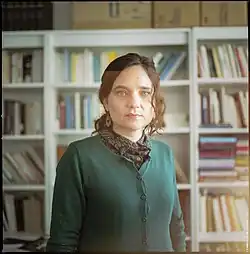 Portrait photograph of a women with brown hair and a green sweater standing in front of a bookshelf filled with books.