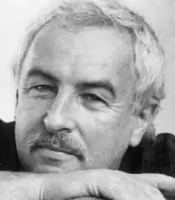 Black and white publicity headshot of a half-smiling, white-haired man with moustache, resting his chin on his hands.