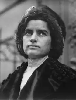 A middle-aged woman wearing a cape and fur hat looks up and to her left.