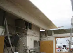 The damaged roof and walls of a hospital, seem from inside. The outside is visible, and mangled metal is piled up in an inlet.