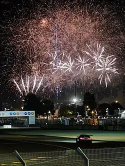 Colour photograph showing a view across a racetrack corner. A BMW M4, silhouetted with red rear lights visible, drives away from the camera. A large fireworks display fills the top half of the photo.