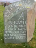 A carved stone with the inscription "Love looks not with the eyes but with the mind / A beloved son, brother & uncle taken from our lives but never from our hearts"