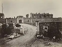 Photograph of the fort of Vanvres, destroyed by the war. In the foreground, a road leading up to it, ending in a bridge. It crosses a low wall which is partly destroyed. In the second plan, the fort's wall and an entrance, located behind a ditch and only slightly damaged. Behind the wall, a few buildings can be seen, including a large barracks of which only parts of the facades remain.