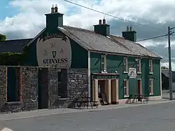 Photo of Linnane's Pub - A green colour building with "Linnane's Pub" written in light yellow above the main door. The door and window frames are light yellow. There is a large advertisement for Guinness on the side of the building. There are two park benches in front of the pub. There is a grey structure on the left side of the pub. The photo was taken during the day when the sky was blue with white clouds. It is taken from outside, and from opposite side of the road