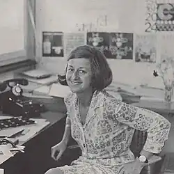 Lora Lamm centered, sitting on a chair, smiling, looking at the camera, sketches on the wall behind her and on the table beside her.