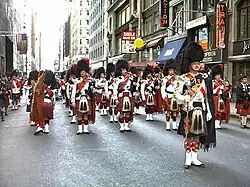 Red-kilted bagpipers in feather bonnets on an urban city street