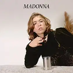 Cover image of "Madonna": a woman wearing a black shirt poses with a tin can in front of her; her middle and ring fingers are covered in a brown sauce. The name of the song in all caps, "MADONNA", is printed above her.