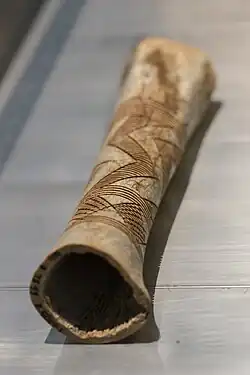 A roughly cylindrical, hollow bone. Zigzagging inter-crossing patterns have been scored into the surface and are highlighted in darker colour.