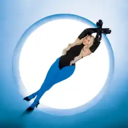 A woman with waist-length bleached hair in tight-fitting blue and black crossing her legs and hands above her head, with a white circle cut out of the blue behind her