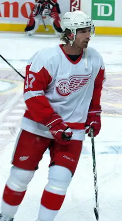 A Caucasian ice hockey player in his thirties. He is skating relaxed on the ice while looking to his right. He wears a white and red jersey, along with a white-visored helmet.