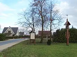 Image of a green lawn next to a street with a house in the background. On the lawn are three trees and a sign in front of the trees.