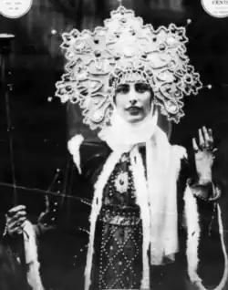 A young white woman with dark eyes, wearing a large Russian-inspired headpieces and robes, one hand raised to show rings on her fingers