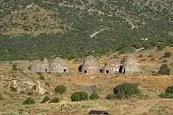 An image depicting 5 brick kilns with small entrances, two of the kilins have collapsed. It is surround by arid vegetation and mountains.
