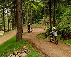 A mother and young child ride mountain bikes on a dirt trail through the forest at Mt. Abram Bike Park. The trail curves gently, and both riders wear helmets.