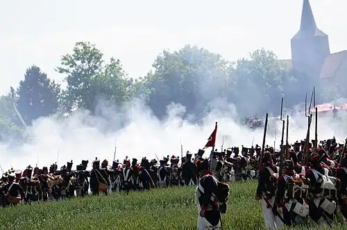 Photo shows a re-enactment of a Napoleonic battle. A French firing line is seen from the rear while a church tower looms to the right.