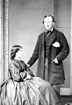 A black and white photo of George Hunter Cary standing next to his wife, seated. Cary stands with his left hand partially in his jacket's pocket, while the other braces his wife's chair. His wife, wearing a large dress which almost completely obscures the chair and Cary's legs, holds her hands on her lap and looks to the right